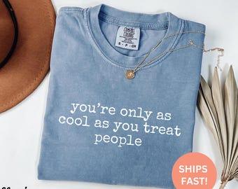 以下が含まれることがあります： 「you're only as cool as you treat people」の白文字がプリントされた、ブルーデニムのComfort Colors Tシャツ。ペンダント付きのゴールドネックレスがシャツの上にかけられています。背景には茶色の帽子があります。