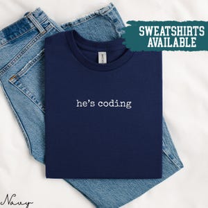 Peut inclure: T-shirt bleu marine plié avec l'inscription blanche "he's coding". Le t-shirt est posé sur un jean bleu. Un rectangle turquoise affiche les mots "SWEATSHIRTS AVAILABLE".