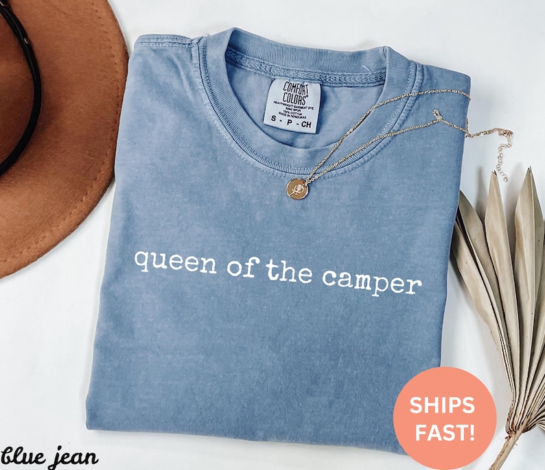 May include: A blue jean Comfort Colors t-shirt with the text "queen of the camper" in white. A gold necklace with a pendant is draped across the shirt. A brown hat is visible in the background.