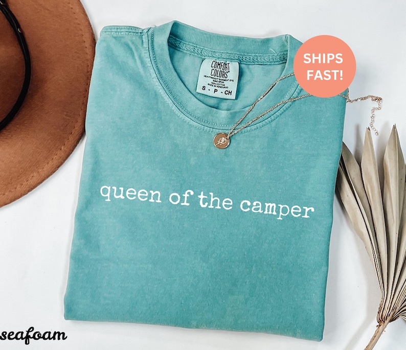 May include: Seafoam green t-shirt with the text "queen of the camper" in white. A gold necklace with a pendant rests on the shirt. The shirt is folded and has a Comfort Colors tag.