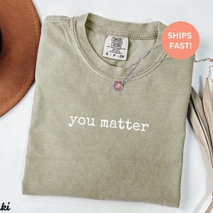 Pode incluir: Camiseta Comfort Colors c&aacute;qui com as palavras "you matter" em branco. Um colar de prata com um pingente rosa repousa sobre a camisa. A camisa est&aacute; dobrada sobre uma superf&iacute;cie branca. A etiqueta diz S-P-CH.