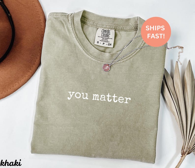 Pode incluir: Camiseta Comfort Colors c&aacute;qui com as palavras "you matter" em branco. Um colar de prata com um pingente rosa repousa sobre a camisa. A camisa est&aacute; dobrada sobre uma superf&iacute;cie branca. A etiqueta diz S-P-CH.