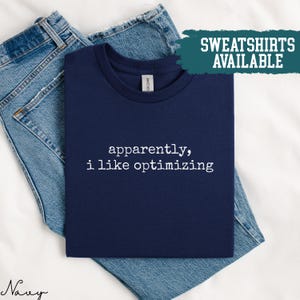 Puede incluir: Camiseta azul marino con el texto "apparently, i like optimizing" en blanco. La camiseta está doblada sobre unos vaqueros azules. Una pancarta turquesa en la esquina superior derecha dice "SWEATSHIRTS AVAILABLE".