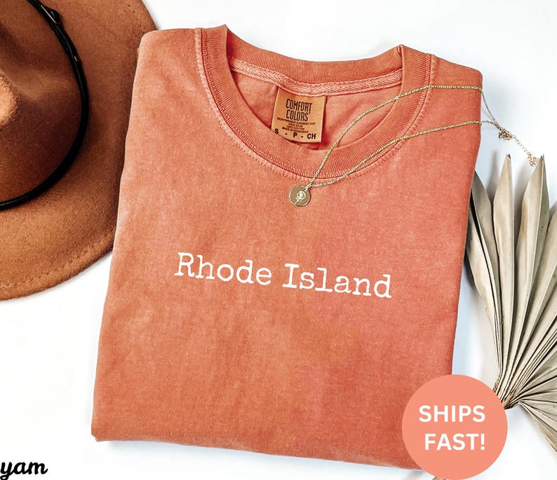 Puede incluir: Una camiseta de color coral con las palabras "Rhode Island" impresas en blanco. La camiseta tiene un cuello redondo y una peque&ntilde;a etiqueta en la parte superior de la espalda. Un collar dorado con un colgante est&aacute; drapeado sobre la camiseta. Un sombrero marr&oacute;n est&aacute; en la esquina superior izquierda.