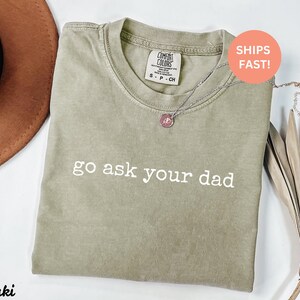 May include: Khaki-colored t-shirt with the phrase "go ask your dad" printed in white. The shirt has a crew neck and short sleeves. A silver necklace with a pendant is draped on the shirt. The shirt is labeled "Comfort Colors".