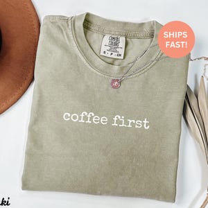 Puede incluir: Camiseta color caqui con las palabras "coffee first" en blanco. La camiseta tiene cuello redondo y una peque&ntilde;a etiqueta en la parte superior de la espalda. Un collar plateado con un colgante est&aacute; sobre la camiseta. Se ve un sombrero marr&oacute;n.