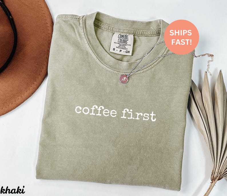 Puede incluir: Camiseta color caqui con las palabras "coffee first" en blanco. La camiseta tiene cuello redondo y una peque&ntilde;a etiqueta en la parte superior de la espalda. Un collar plateado con un colgante est&aacute; sobre la camiseta. Se ve un sombrero marr&oacute;n.