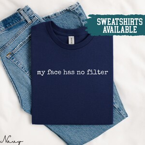 May include: Navy blue t-shirt with the text "my face has no filter" in white. The shirt is folded on top of a pair of blue jeans. The words "SWEATSHIRTS AVAILABLE" are in a teal rectangle.