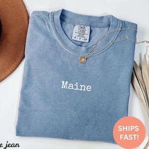 Könnte beinhalten: Ein blaues Comfort Colors T-Shirt mit dem Wort "Maine" in Weiß gestickt. Eine goldene Halskette mit Anhänger liegt auf dem Shirt. Das Shirt ist gefaltet, und ein brauner Hut ist im Hintergrund zu sehen. Das Bild enthält auch einen "SHIPS FAST!" Aufkleber.