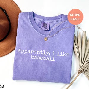 May include: A lilac t-shirt with the text "apparently, i like baseball" in white. A gold necklace with a pendant is on the shirt. A brown hat and dried palm leaf are also in the image.