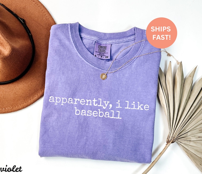May include: A lilac t-shirt with the text "apparently, i like baseball" in white. A gold necklace with a pendant is on the shirt. A brown hat and dried palm leaf are also in the image.
