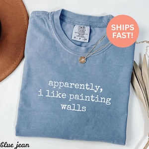 May include: A blue jean Comfort Colors t-shirt with the text "apparently, i like painting walls" in white. The shirt has a crew neck and short sleeves. A gold necklace with a pendant is draped on the shirt.