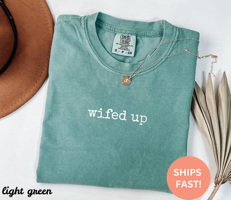 Puede incluir: Una camiseta verde claro con la frase "wifed up" en letras blancas. La camiseta tiene cuello redondo y mangas cortas. Un collar dorado con un colgante est&aacute; drapeado sobre la camiseta. La camiseta est&aacute; doblada sobre una superficie blanca.