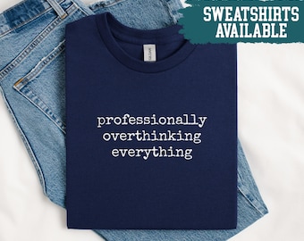 May include: Navy blue t-shirt with the text "professionally overthinking everything" in white. The shirt is folded on top of a pair of blue jeans. A teal banner in the upper right corner reads "SWEATSHIRTS AVAILABLE".