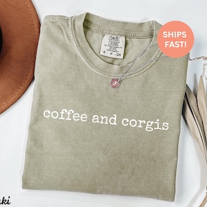 Puede incluir: Camiseta de color caqui con las palabras "coffee and corgis" impresas en blanco. La camiseta tiene cuello redondo y mangas cortas. Un collar plateado con un colgante est&aacute; sobre la camiseta. Un sombrero marr&oacute;n y hojas secas en el fondo.