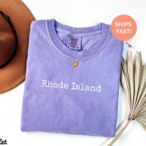 Puede incluir: Una camiseta violeta con las palabras "Rhode Island" impresas en blanco. Un collar de oro con un colgante est&aacute; drapeado sobre la camiseta. Un sombrero marr&oacute;n y una hoja de palma seca est&aacute;n en el fondo.
