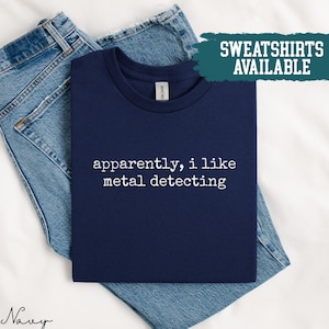 Könnte beinhalten: Marineblaues T-Shirt mit dem weißen Text "apparently, i like metal detecting". Das T-Shirt ist auf einer blauen Jeans gefaltet. Ein türkisfarbenes Rechteck zeigt die Worte "SWEATSHIRTS AVAILABLE".
