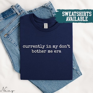 Könnte beinhalten: Marineblaues T-Shirt mit dem weißen Text "currently in my don't bother me era". Das T-Shirt liegt gefaltet auf einer blauen Jeans. Ein türkisfarbenes Rechteck oben rechts zeigt "SWEATSHIRTS AVAILABLE".