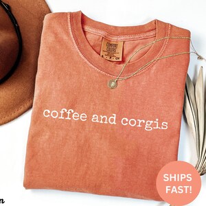 Puede incluir: Una camiseta Comfort Colors de color coral con las palabras "coffee and corgis" en blanco. Un collar dorado y un sombrero marr&oacute;n est&aacute;n al lado de la camiseta. Una pegatina que dice "SHIPS FAST!" est&aacute; en la parte inferior derecha.