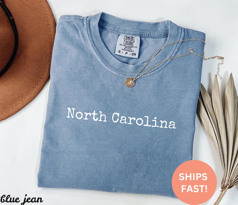 Op de afbeelding: Een blauw Comfort Colors t-shirt met de tekst "North Carolina" in het wit. Een gouden ketting met een hanger is over het shirt gedrapeerd. Een bruine hoed en gedroogde bladeren zijn op de achtergrond.