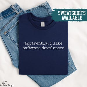 Może przedstawiać: Granatowa koszulka z białym napisem "apparently, i like software developers". Koszulka jest złożona na parze niebieskich dżinsów. Napis "SWEATSHIRTS AVAILABLE" znajduje się na turkusowym banerze.