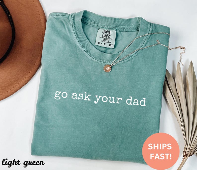 May include: Light green t-shirt with the phrase "go ask your dad" in white text. The shirt has a crew neck and short sleeves. A gold necklace with a pendant is draped over the shirt. The shirt is folded on a white surface.
