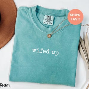 Puede incluir: Camiseta verde mar con la frase "wifed up" en letras blancas. Un collar dorado con un colgante est&aacute; colocado sobre la camiseta. La camiseta est&aacute; doblada y colocada sobre una superficie blanca. Un sombrero marr&oacute;n es visible en el fondo.