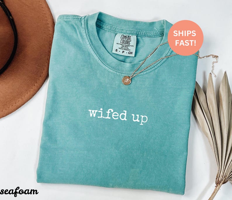 Puede incluir: Camiseta verde mar con la frase "wifed up" en letras blancas. Un collar dorado con un colgante est&aacute; colocado sobre la camiseta. La camiseta est&aacute; doblada y colocada sobre una superficie blanca. Un sombrero marr&oacute;n es visible en el fondo.