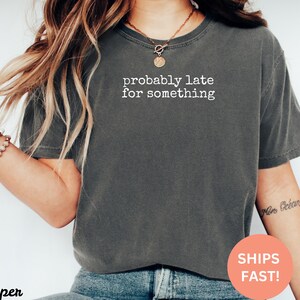 Puede incluir: Camiseta gris oscuro con el texto blanco "probably late for something". La camiseta de estilo casual es adecuada para el uso diario. La imagen tambi&eacute;n incluye una pulsera y un collar.