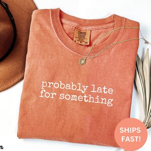 Puede incluir: Una camiseta Comfort Colors de color coral con la frase "probably late for something" en texto blanco. Un collar dorado con un colgante descansa sobre la camiseta. Un sombrero marr&oacute;n es visible en el fondo.