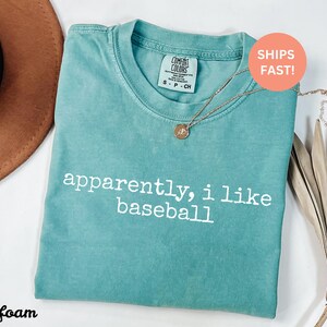 May include: Seafoam green t-shirt with the text "apparently, i like baseball" in white. The shirt has a crew neck and a small tag on the collar. A gold necklace with a pendant is draped on the shirt.