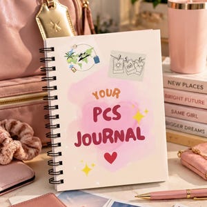 May include: A spiral-bound journal with a cream cover featuring a pink watercolor heart, yellow star accents, and the text "YOUR PCS JOURNAL" in red. The cover includes illustrations of a world map, travel icons, and hanging polaroid photos.