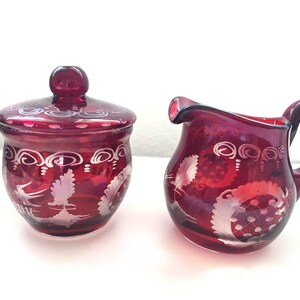 May include: A deep red glass sugar bowl with a matching lid and a matching creamer jug. Both pieces feature etched white floral and bird motifs around the exterior, with a smooth, translucent finish and a rounded, bulbous shape.