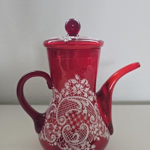 Peut inclure: Une théière en verre rouge rubis translucide ornée d'un délicat motif de dentelle peint en blanc sur le corps et le couvercle. La théière présente une anse incurvée, un long bec effilé et un bouton rond sur le couvercle. Le verre est lisse.