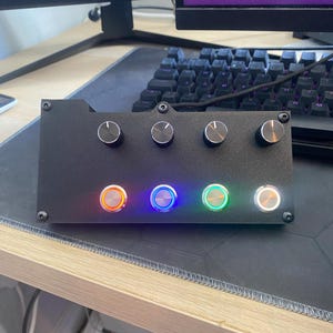 May include: Black rectangular control panel with four knobs and four illuminated buttons. The buttons glow orange, blue, green, and white. The panel is mounted with four screws and sits on a desk near a keyboard and monitor.