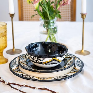 May include: A black and white ceramic bowl with a yellow and white geometric design sits on a black and white ceramic plate with a yellow and white geometric design. The plate is on a larger black and white ceramic plate with a yellow and white geometric design. The plates are on a white tablecloth.