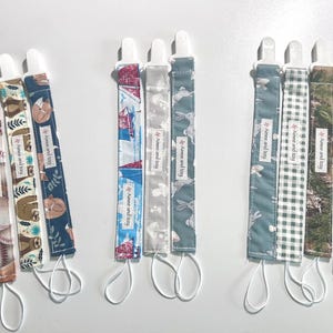 Puede incluir: Una colección de clips para chupetes con varios patrones de tela. Los diseños incluyen béisboles, perezosos, zorros, conejos, cuadros vichy e estampados florales. Cada clip tiene un clip de plástico blanco y un lazo. El texto "by Anne and Izzy" está presente.