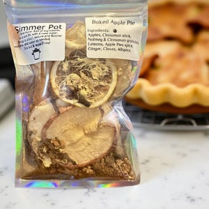 以下が含まれることがあります： 「Simmer Pot」と書かれた透明なポーチには、乾燥リンゴのスライス、シナモンスティック、スパイスが入っています。「Baked Apple Pie」の材料として、リンゴ、シナモン、ナツメグ、レモンなどがラベルに記載されています。焼き上がったパイが背景に一部写っています。