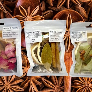 May include: Four small clear plastic bags filled with various dried ingredients for making simmer pot drinks. The bags are labeled "Simmer Pot" and include the names "Warm & Fuzzy", "Chai Latte", and "Lavender Fields". The bags are arranged on a background of star anise.