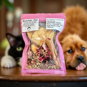 May include: A pink bag labeled "Simmer Pot" filled with dried ingredients including chamomile, apples, pears, cinnamon sticks, rose petals, and hibiscus. The bag is in front of a black and white cat and a brown dog.
