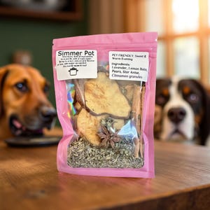 Peut inclure: Un sachet rose de "Simmer Pot" pour animaux de compagnie, contenant de la lavande, de la mélisse, des poires, de l'anis étoilé et des granulés de cannelle. Le sachet est sur une table en bois, avec deux chiens en arrière-plan.