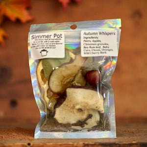 Puede incluir: Una bolsa holográfica transparente etiquetada como "Simmer Pot" que contiene peras secas, manzanas, gránulos de canela, hojas de laurel, clavos, naranjas y corteza de cerezo silvestre. La bolsa está sellada y lista para usar.