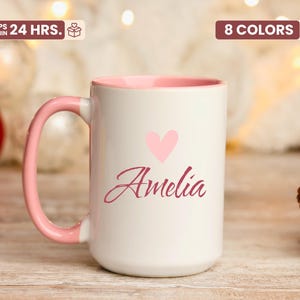 May include: White ceramic mug with a pink interior and handle. The mug features a pink heart and the name "Amelia" in a cursive font. The mug is a gift item.