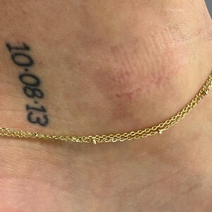 May include: A gold chain anklet with small beads on a person's ankle. The ankle has a tattoo that says "10-08-13".