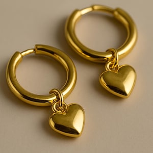 Puede incluir: Un par de pendientes de aro dorados. Cada pendiente presenta un pequeño dije en forma de corazón colgando del aro. Los pendientes tienen una superficie pulida y reflectante, sobre un fondo neutro. Joyería.