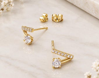 May include: A pair of gold stud earrings featuring a V-shaped bar set with small clear stones and a single larger round clear stone at the base. The earrings include matching gold butterfly backings for secure fastening.