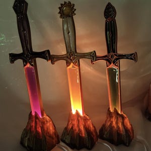 Könnte beinhalten: Drei schwertförmige Lampen auf felsigen Sockeln. Das linke Schwert leuchtet violett, das mittlere orange und das rechte grün. Jedes Schwert hat ein eigenes Griffdesign, darunter Knäufe in Form einer Spitze, einer Sonne und eines Blattes.