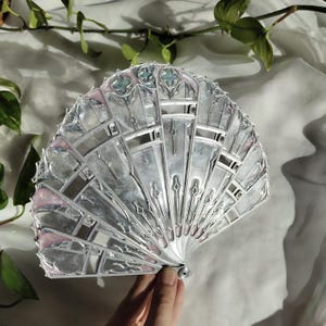 May include: A folding hand fan featuring a silver frame with intricate cutouts and structural ribs. The fan panels are decorated with a shimmering silver finish, accented by soft pink and light blue details along the top and side edges.