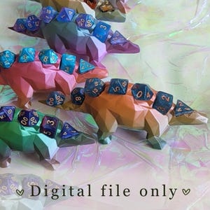 Puede incluir: Tres coloridos soportes para dados con forma de dinosaurio, cada uno de un color diferente: verde, rosa y naranja. Cada dinosaurio tiene un conjunto de dados poliédricos en su espalda. El texto "Digital file only" está en la parte inferior.