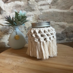 May include: A glass jar wrapped in a cream-coloured macrame cord cover featuring a knotted lattice pattern and a fringed bottom edge. The jar has a silver-coloured metal screw-top lid. The macrame design is intricate with repeating knots.
